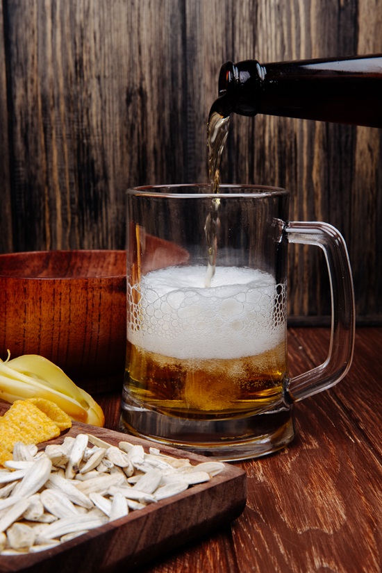 side view of various salty beer snacks on a wooden platter and pouring beer into a mug on rustic wooden background
